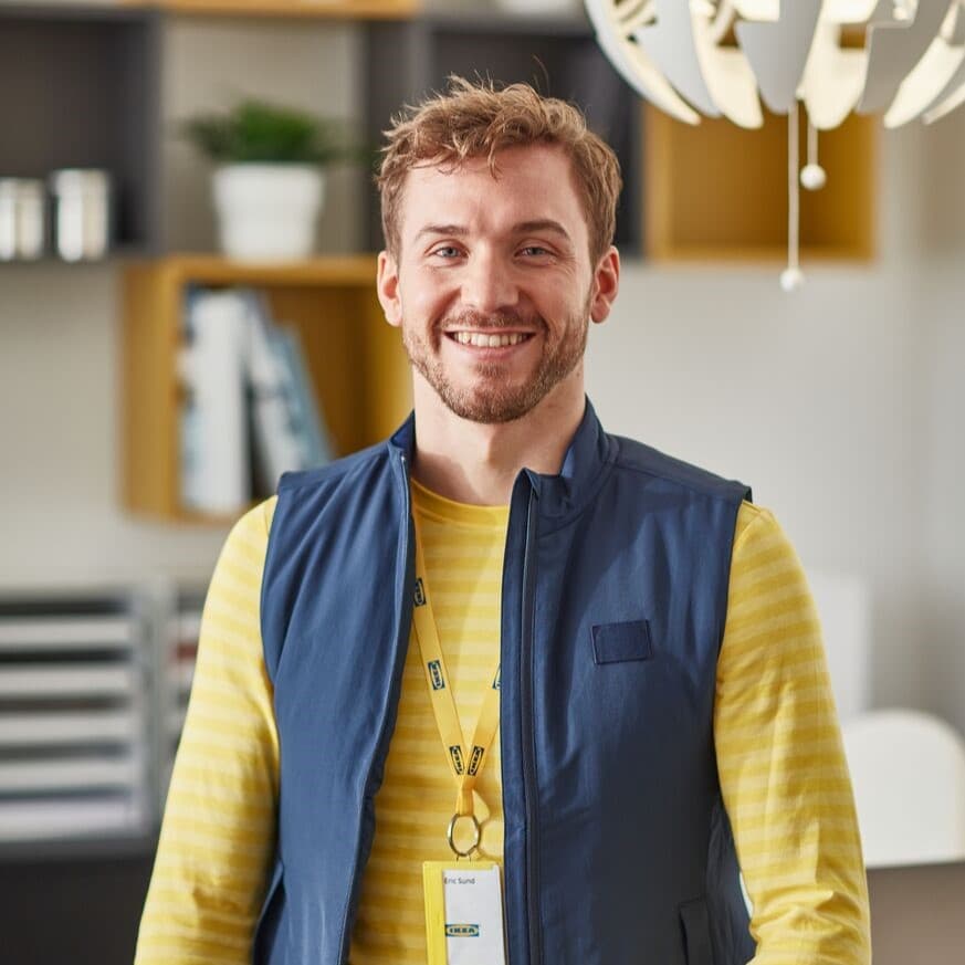 Empleat d’IKEA somrient, amb armilla blava i samarreta groga de ratlles, en un entorn d’oficina amb prestatgeries i una làmpada decorativa al fons.