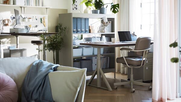 En arbetsplats hemma i neutrala färger i hörnet av ett vardagsrum med två IDÅSEN sitt-/ståbord placerade i olika höjder.