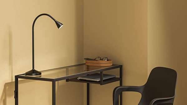 En arbetsstation med ett svartbrunt skrivbord i härdat glas och metall. På bordet står en svart NÄVLINGE-bordslampa samt en skål med glasögon. En svart kontorsstol är placerad bredvid skrivbordet, och rummet har ljusgula väggar.