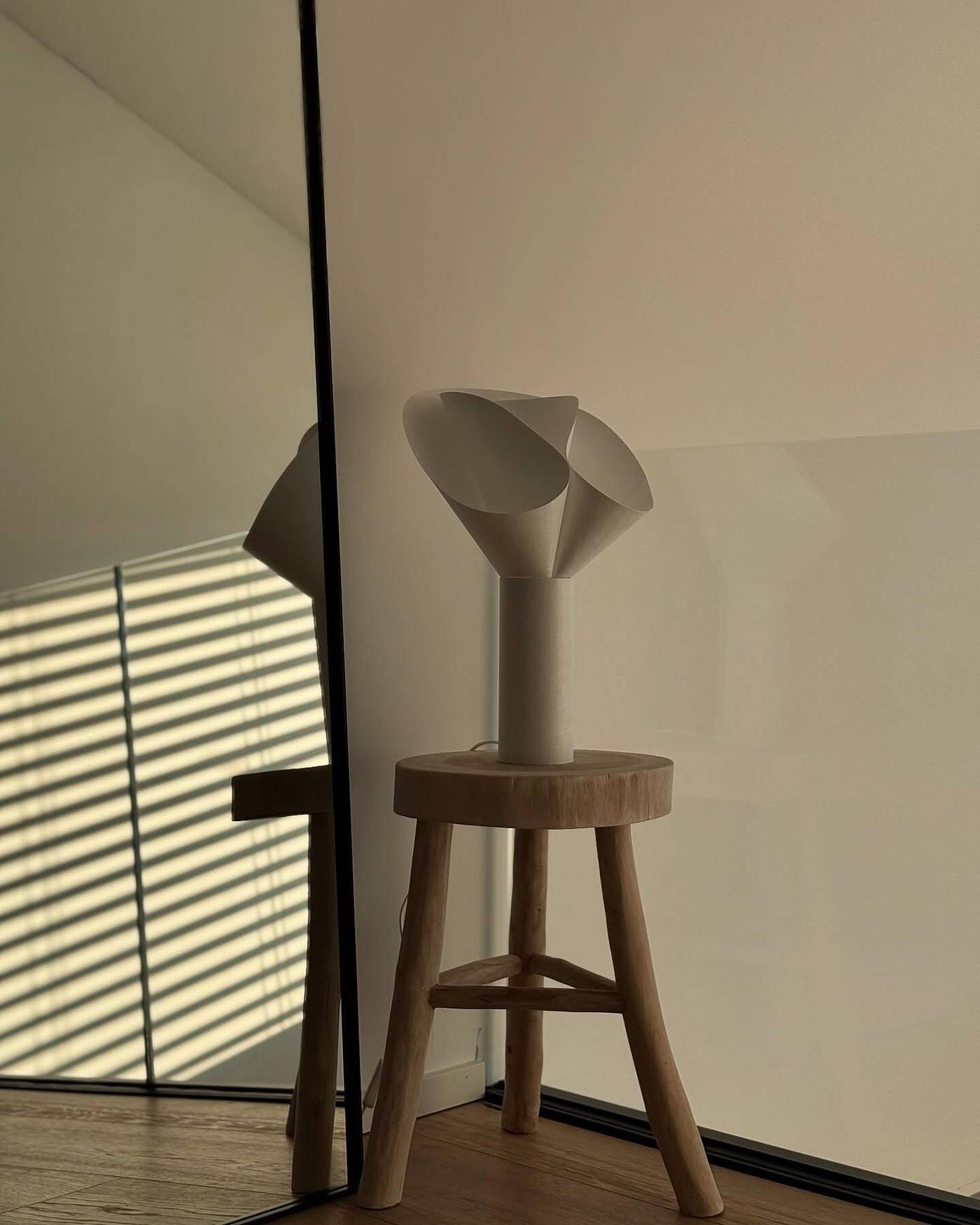 En BÄCKNATE bordslampa står på en träpall framför en stor spegel i ett rum med mjukt ljus och skuggor från fönsterpersienner.