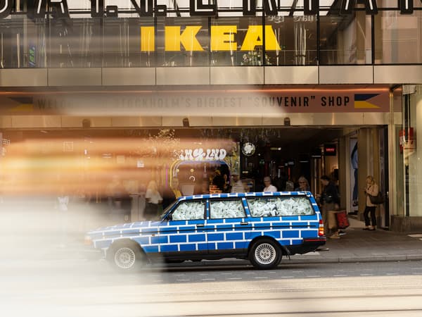 En bil med ett blåvitt rutmönster står parkerad framför ett IKEA-varuhus. Bilen är fylld med vita vattenkannor.