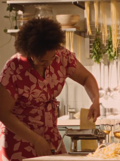 En bild på en person som gör egen pasta i ett kök. Personen är iklädd en rödblommig klänning och matar en pastadeg genom en pastamaskin. I bakgrunden hänger färsk pasta och örter på tork.
