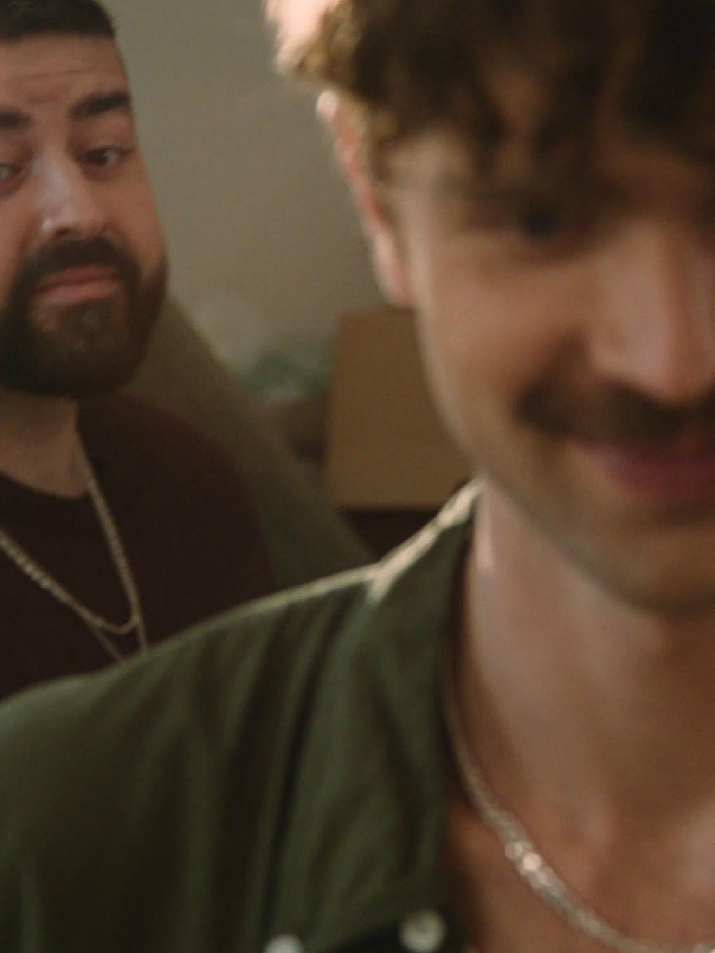 En bild på två män. En man med lockigt hår och mustasch är i förgrunden, delvis ur fokus, medan en skäggig man i bakgrunden tittar på honom med ett observerande uttryck.