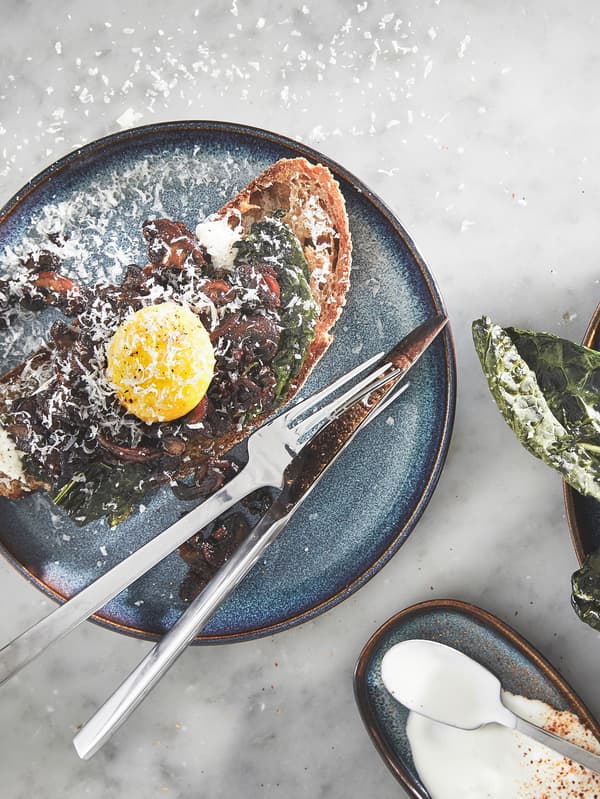 En blå GLADELIG tallerken med et stykke højtbelagt smørrebrød samt en gaffel og en kniv placeret ved siden af.