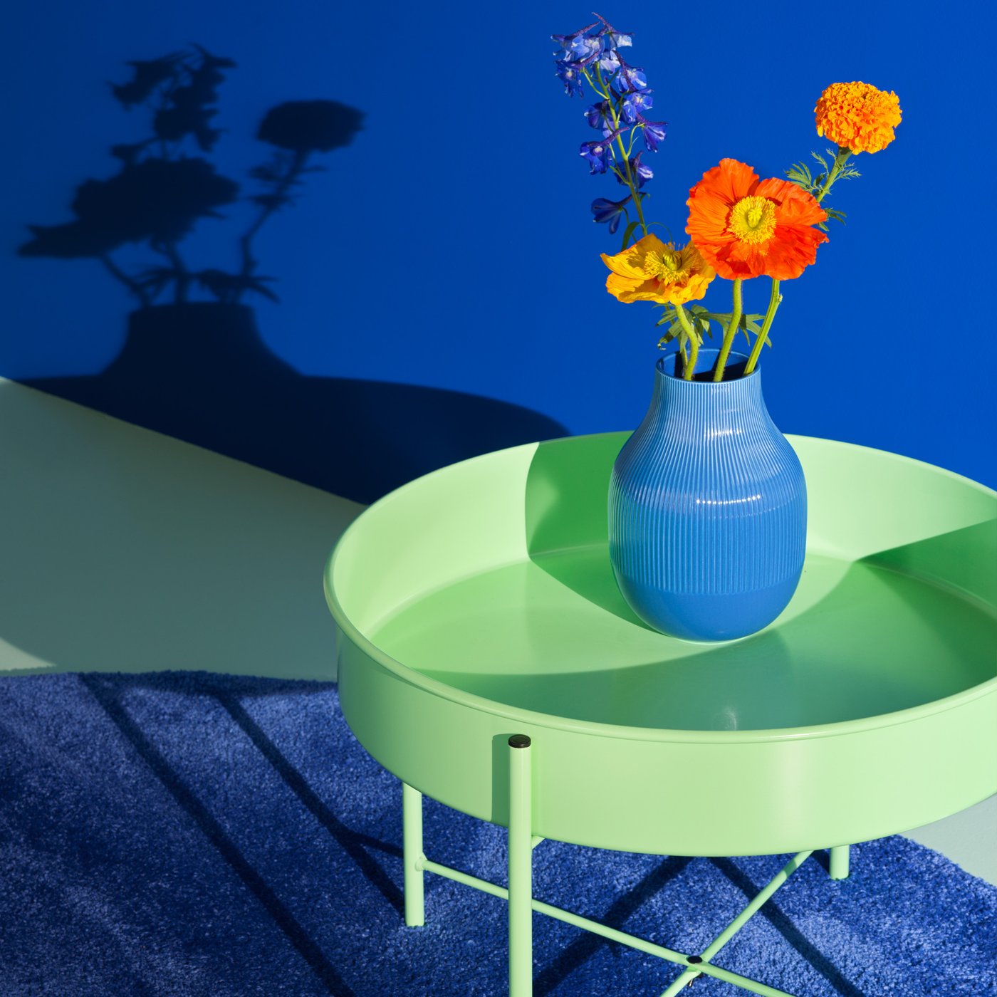 En blå vase med orange blomster i står på et lysegrønt NYTILVERKAD  rundt sidebord som står på et blåt gulvtæppe og et lysegrønt gulv blomsterne kaster skygger op på den blå væg.