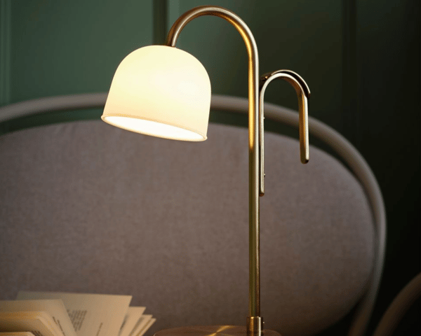En bordslampa i mässingsfärg och vit glasskärm står mot en grå och grön bakgrund.