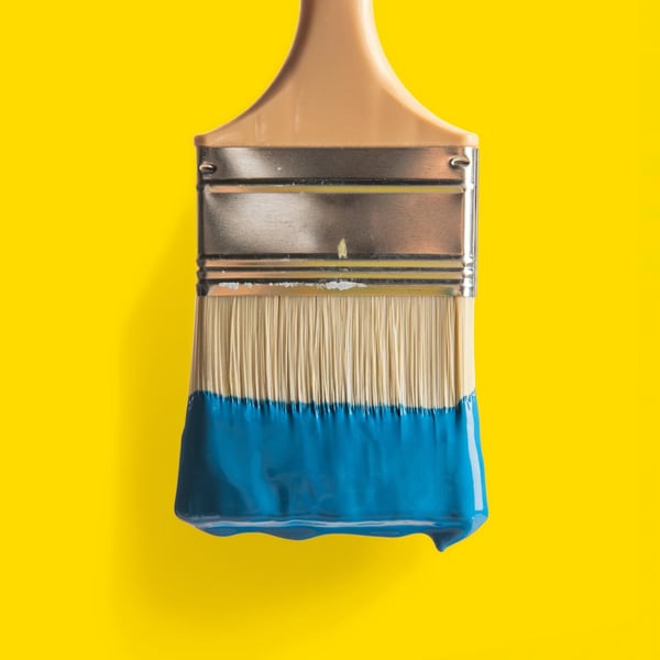 En bred pensel med trähandtag och metallfäste, där borsttopparna är doppade i blå färg, mot gul bakgrund.