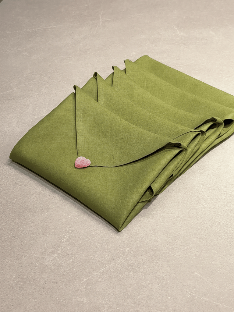 En bunt gröna, vikta linneservetter som ligger på en ljusgrå stenbänk, med ett litet rosa hjärtformat godis placerat på den översta servetten.