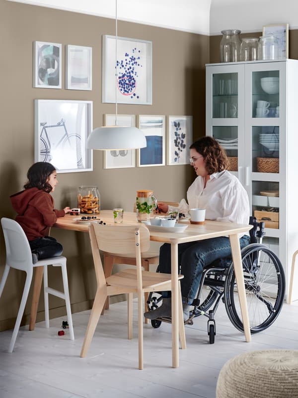 En dame sidder i en kørestol og spiser morgenmad med en pige der knæler på en anden stol ved bordet.