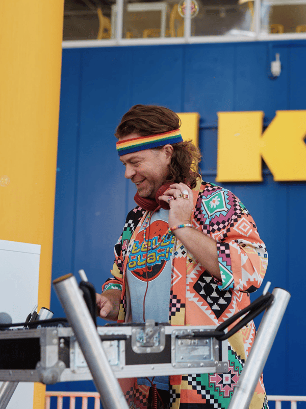 En DJ med et regnbuefarvet pandebånd og en spraglet vest spiller på en mixerpult foran et gult og blåt IKEA logo