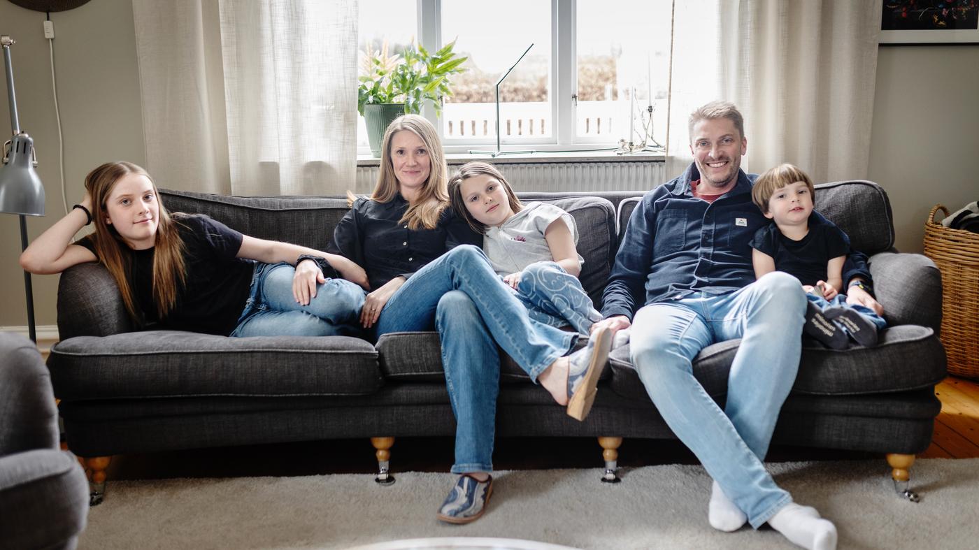 En familj på fem personer, två vuxna och tre barn, sitter i en soffa i ett vardagsrum. 