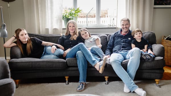 En familj på fem personer, två vuxna och tre barn, sitter i en soffa i ett vardagsrum. 