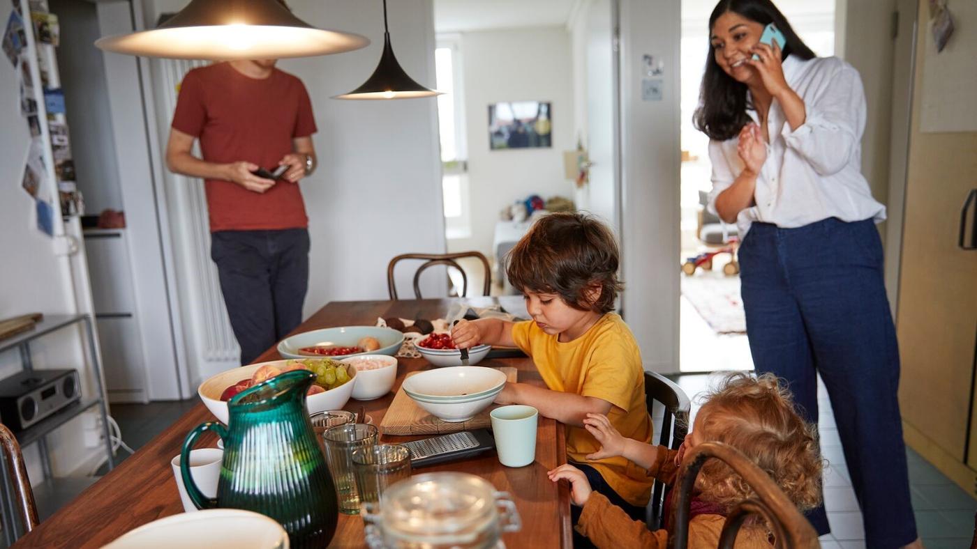 En familj vid matbordet, två barn sitter och en vuxen står i närheten, en annan vuxen kommer in.