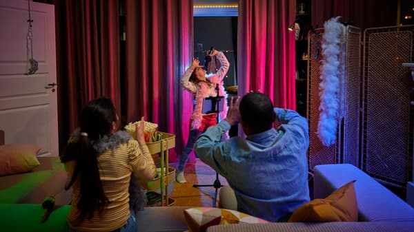 En flicka står framför en soffa och sjunger. I handen har hon en mikrofon. I soffan sitter två vuxna. 