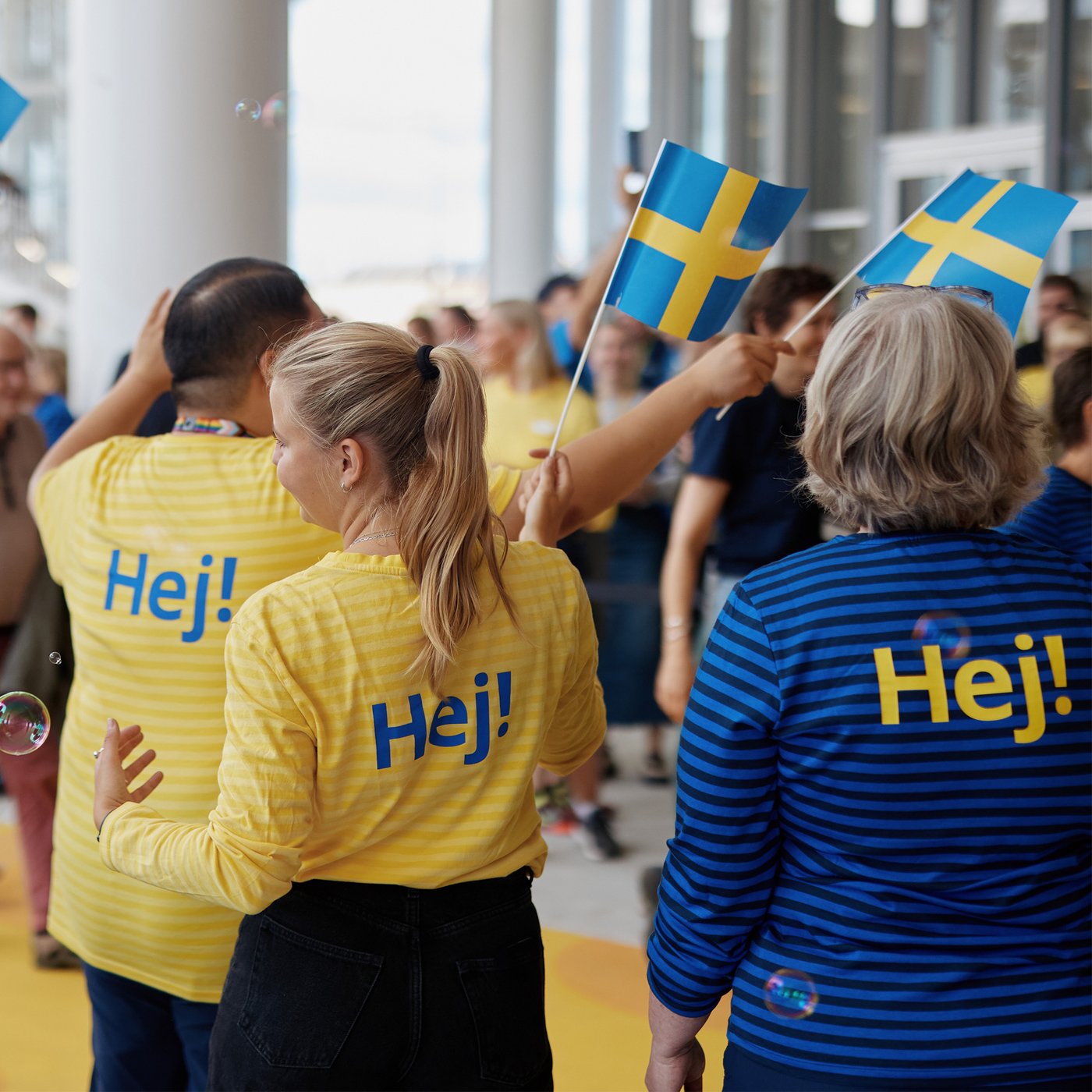 En flok IKEA medarbejdere med gule trøjer hvor der står Hej med blå skrift står på en gul løber og flager til åbningen af IKEA København med det svenske flag.