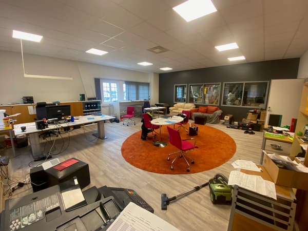 En före-bild som visar ett stökigt kontor före ett inredningsuppdrag. I mitten av rummet finns en rund orange matta och ett bord med stolar, och längs väggarna syns förvaring, skrivare, soffor och fler bord.
