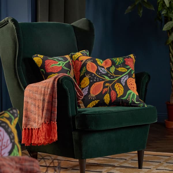 En grön fåtölj med två färgglada,mönstrade kuddar i och en orange pläd hängandes på armstödet står på en gul matta.