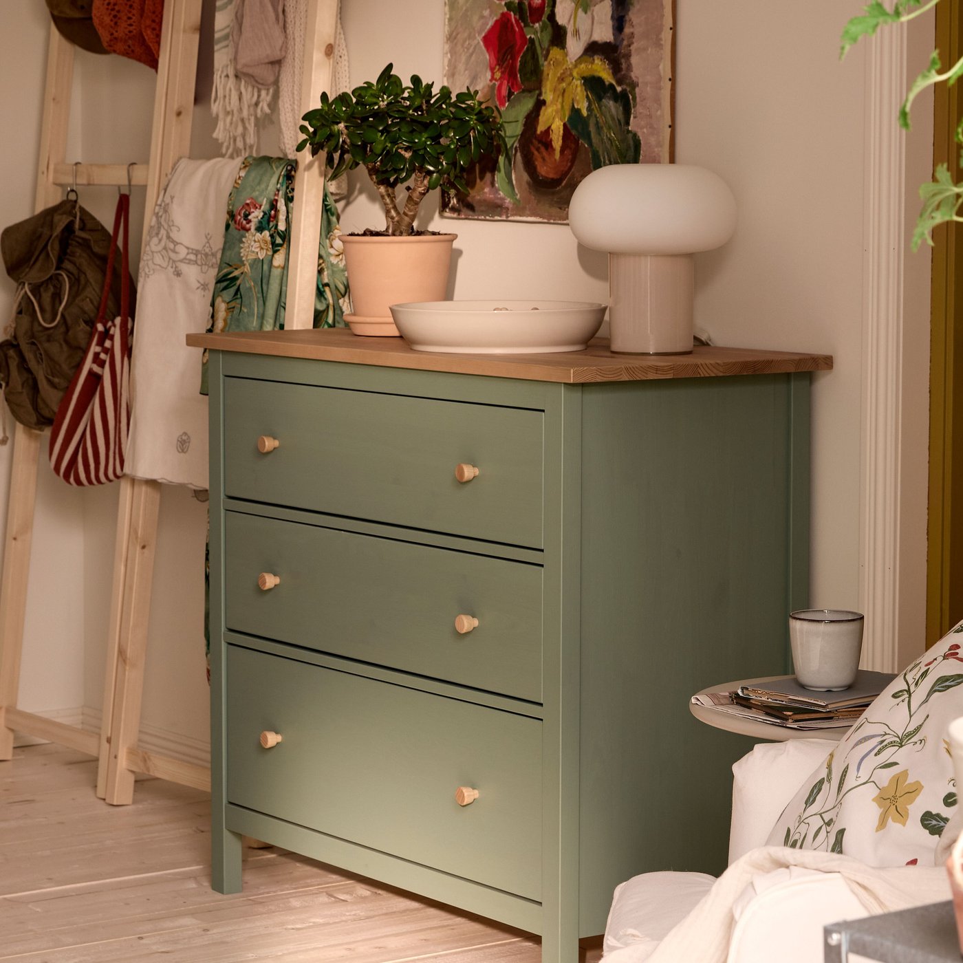 En grön HEMNES byrå med trätopp står i ett rum, dekorerad med en bordslampa, ett fat och en krukväxt. Bredvid syns en trästege med tyg och väskor.
