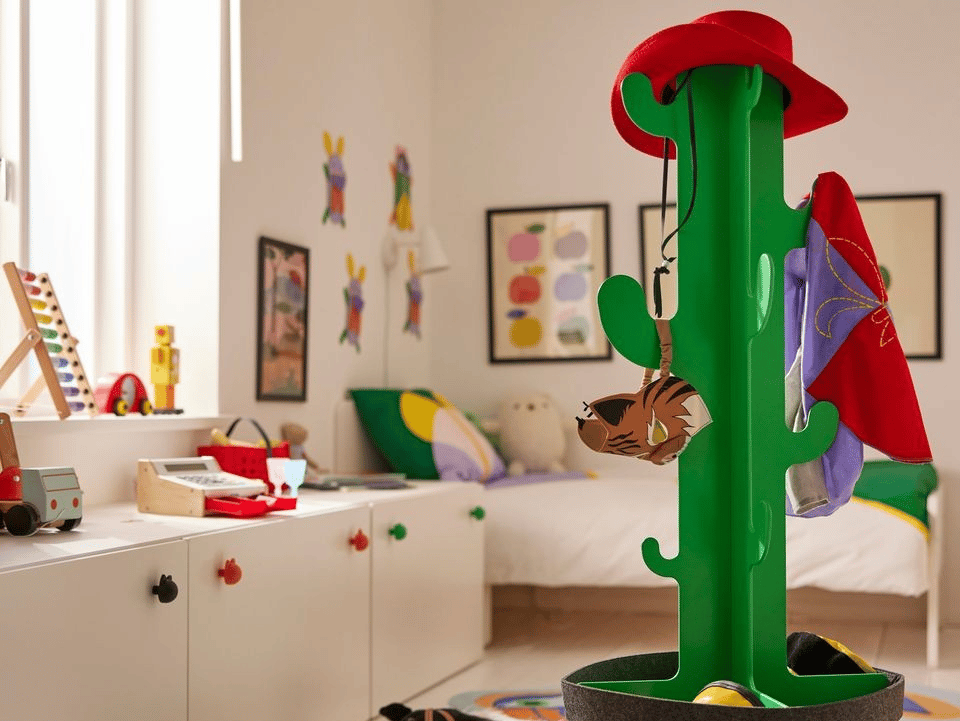 En grön klädhängare i form av en kaktus står i ett vitmålat, ljust rum. På klädhängaren hänger en röd cowboyhatt, en djurmask och en lila och röd pläd.