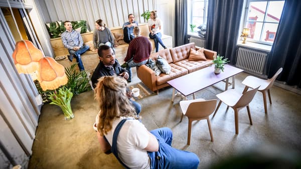 En grupp människor i ett ljust rum med modern inredning. Rummet har en soffa, flera stolar, ett soffbord med växter samt stora fönster med gardiner. Det finns även hängande lampor och fler växter runtom i rummet.