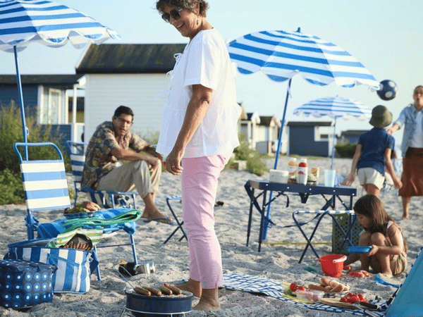 En grupp människor på stranden med parasoll, stolar och en grill. Barn leker i sanden.