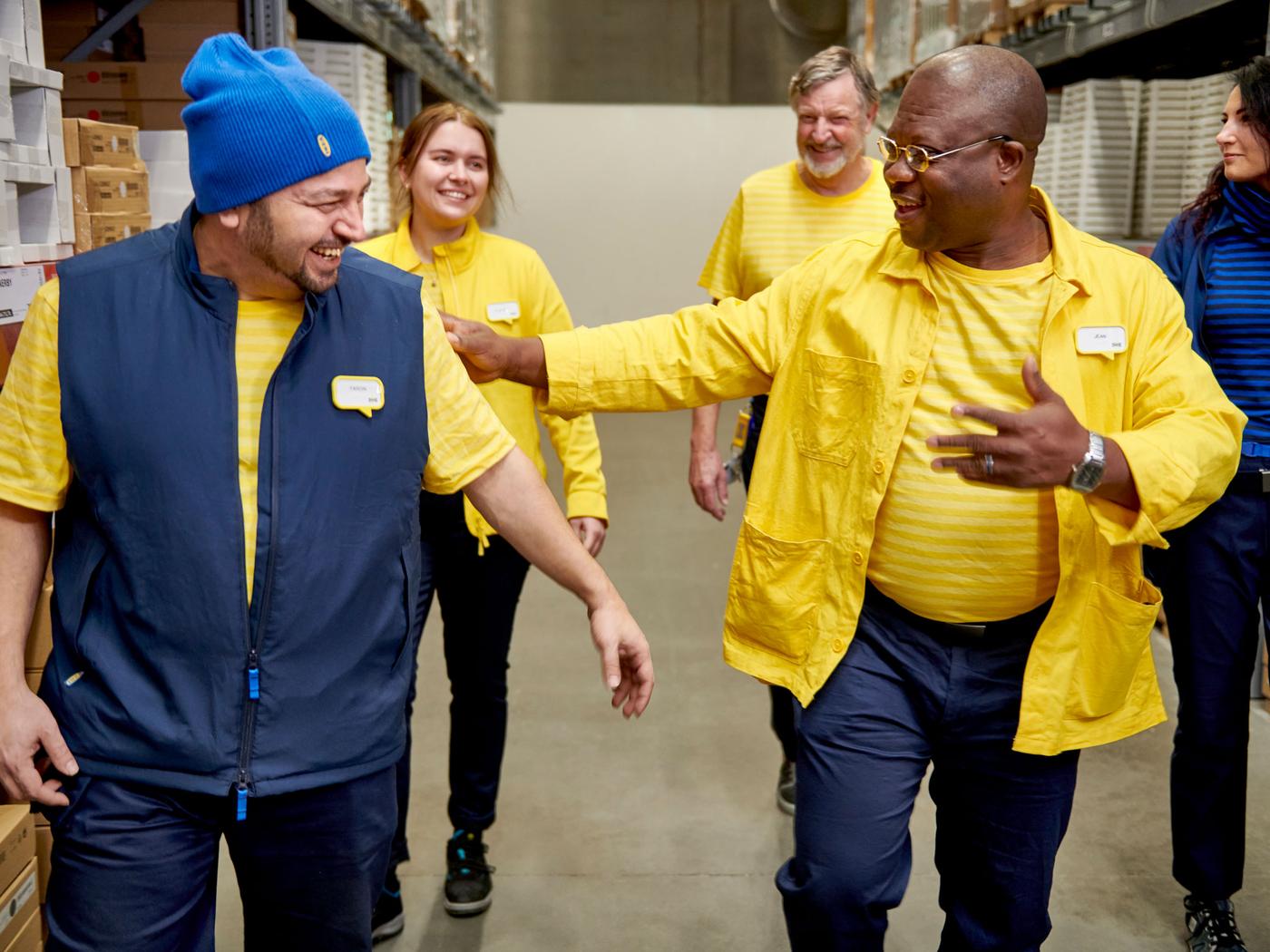 En gruppe medarbejdere i et lager går sammen og griner. Forrest ses to mænd, begge iført gule trøjer og mørkeblå veste eller jakker. Den ene mand bærer en blå hue og smiler bredt, mens den anden, i en gul jakke og briller, griner og klapper ham venligt på skulderen. Bag dem går flere kolleger, alle iført lignende gule og blå uniformer. Omgivelserne er et lager med hylder fyldt med kasser og produkter. Stemningen er afslappet og venlig, som om de nyder deres arbejde og samvær.