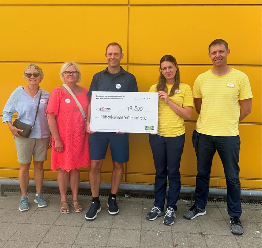 En gruppe mennesker står foran en gul væg mens de holder en skilt hvor der står at IKEA har doneret penge til Børns Voksenvenner.
