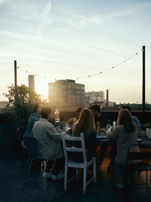 En gruppe personer sitter på stoler rundt et bord. De drikker og spiser i solnedgangen over bygningene i bakgrunnen.
