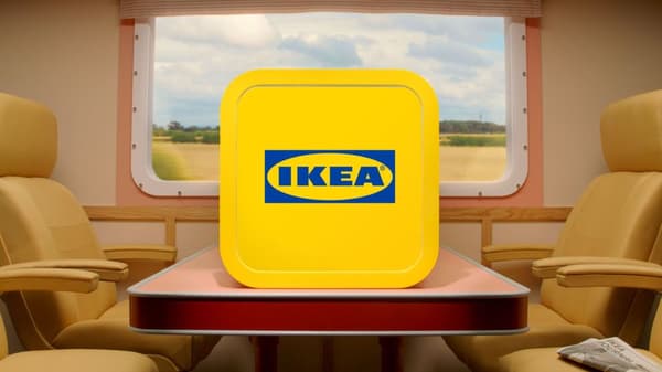 En gul box (appikon) med en IKEA logga står placerad på ett bord mellan två rader säten i ett tåg