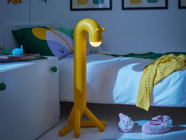 En gul golvlampa i form av en giraff står vid sängen i ett barnrum som är halvdunkelt. På den vita sängen ligger en gul pläd och på mattan står rosa morgontofflor.