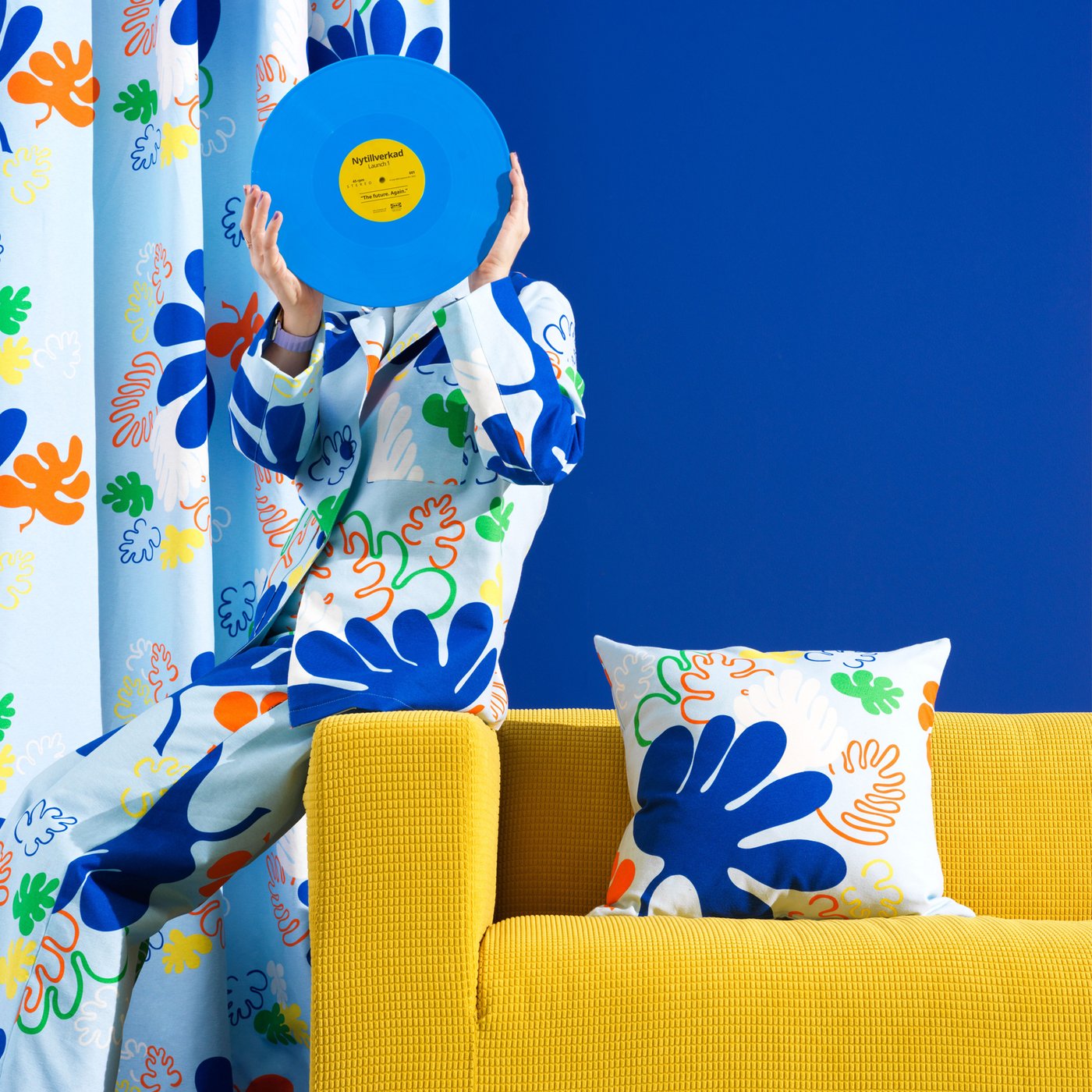 En gul sofa med en blomstret pude med NYTILVERKAD pudebetræk bag sofaen hænger et NYTILVERKAD gardin og en dreng står i en NYTILVERKAD pyjamas og skjuler sit ansigt med en blå og gul plade.