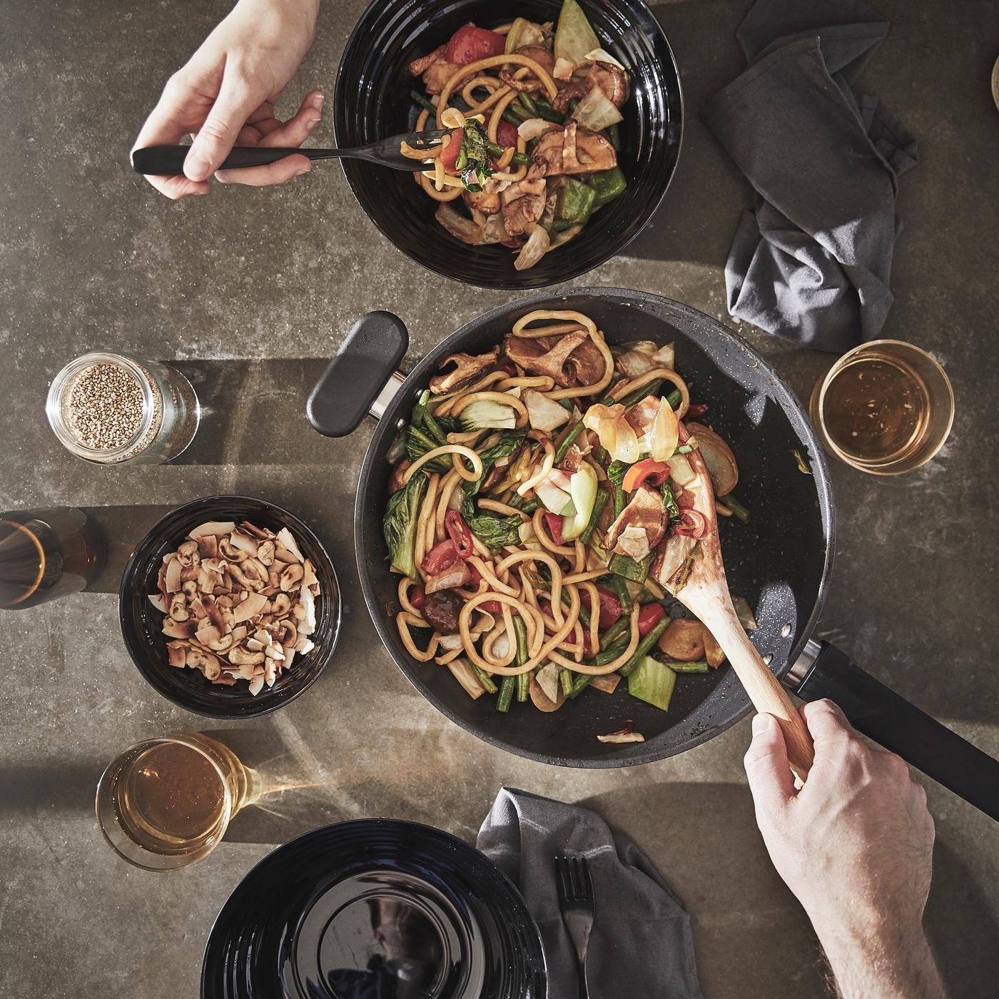 En hånd bruker en tresleiv til å servere en grønnsaks- og nudelrett fra en MIDDAGMAT-wok over i to svarte boller ved siden av.