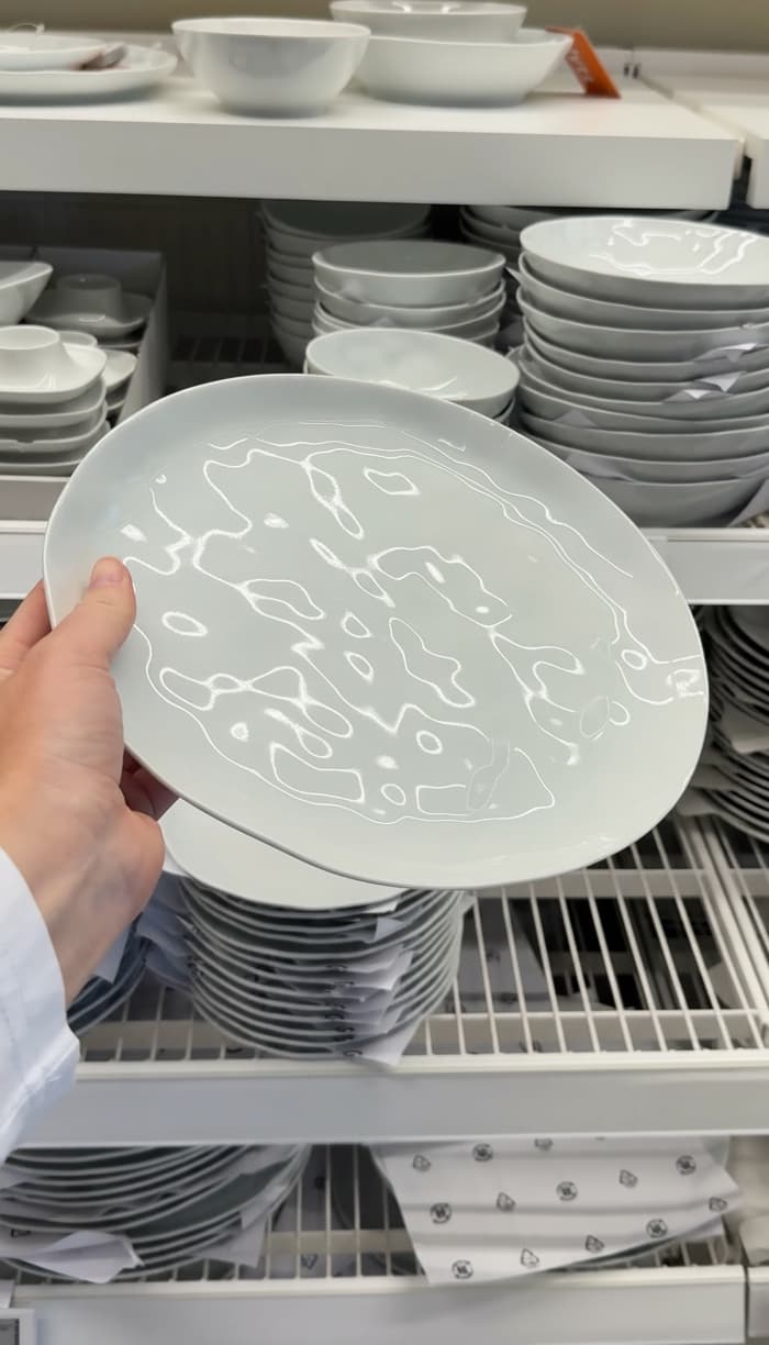 En hand håller en vit tallrik framför hyllor fyllda med staplade, vita porslinstallrikar och skålar i ett varuhus.
