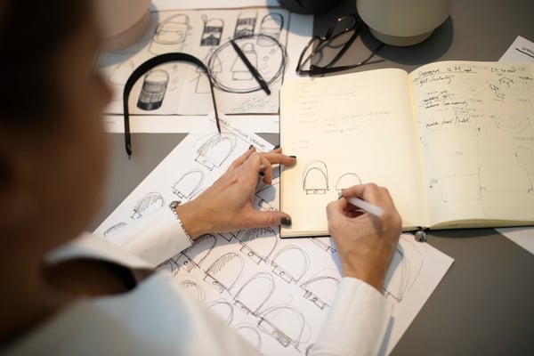 En hand ritar skisser i ett block som ligger ovan andra skisser som är ritade på ett papper