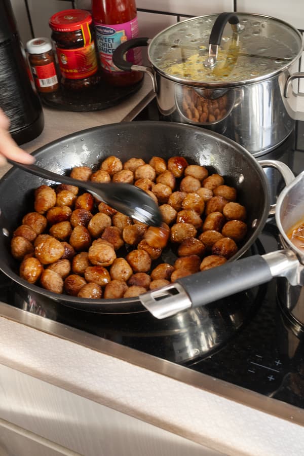 En hand rör i en stekpanna med köttbullar som står på spisen. Där står också en gryta med potatis och en gryta med brunsås. 