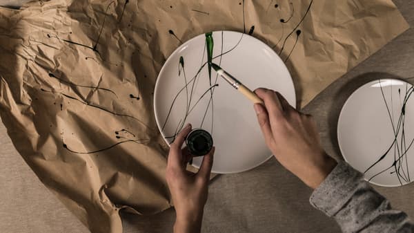 En hand som stänker grön porslinsfärg med pensel på IKEA FLITIGHET vit tallrik.