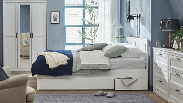 En hvid seng med opbevaring under står i et soveværelse med en kommode med seks skuffer i samme stil samt et garderobeskab i fra samme serie.