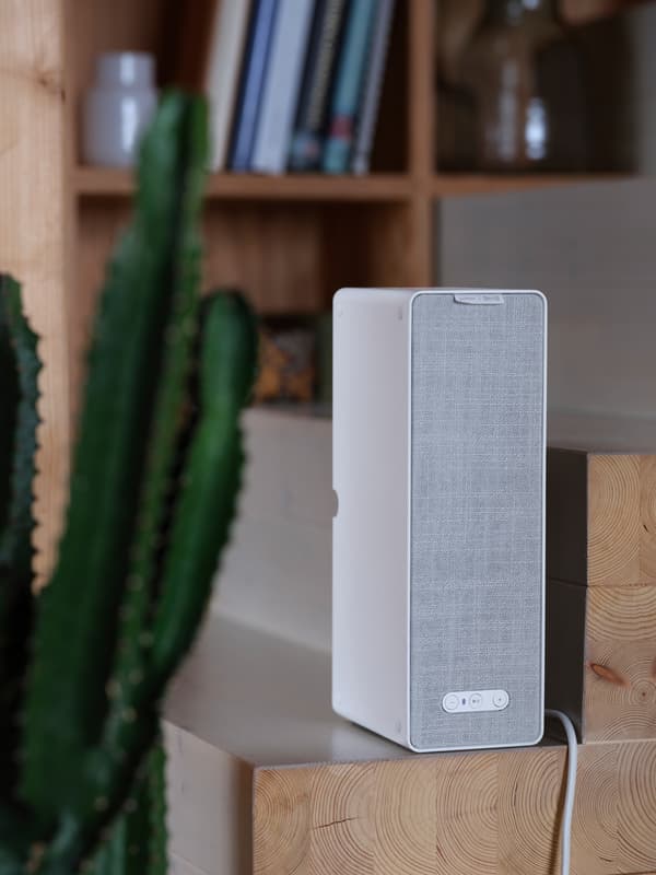 En hvit SYMFONISK trådløs bokhyllehøyttaler står i ei trapp av tre med ei bokhylle i bakgrunnen.