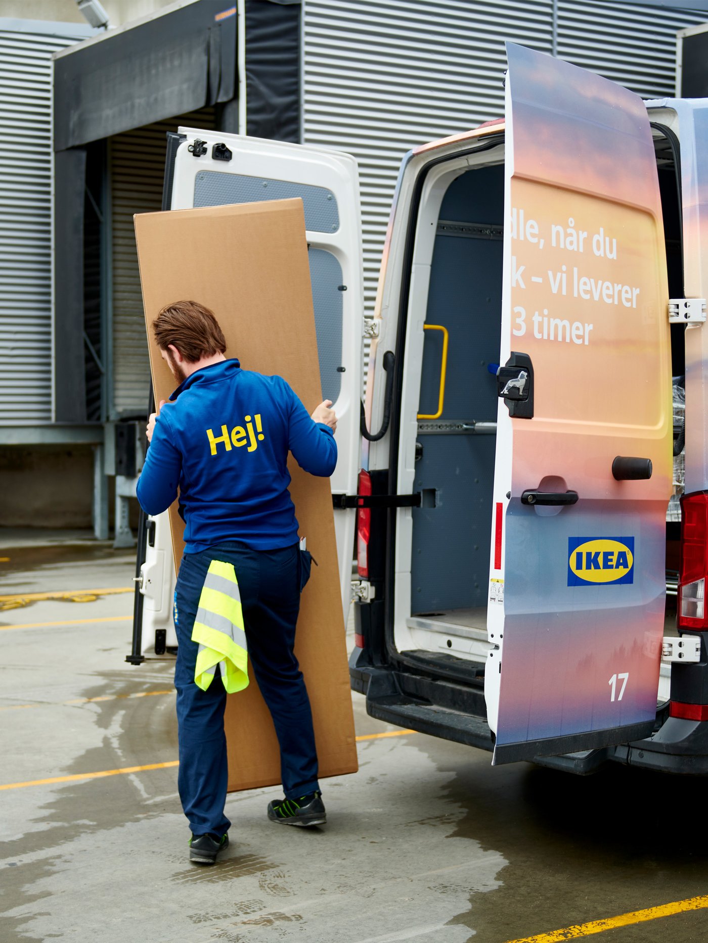 En IKEA medarbejder er ved at løfte en aflang pakke ud af en IKEA varevogn med en åben bagsmæk og ind i en lagerbygning.