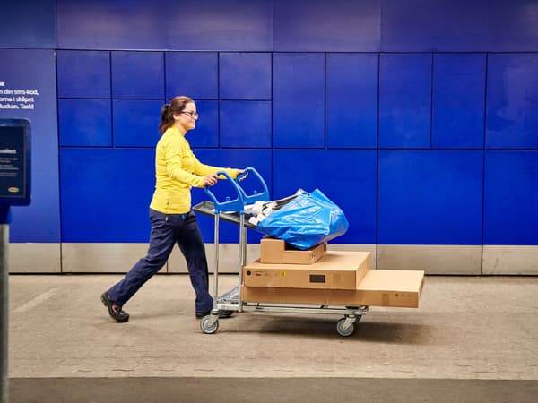 En IKEA medarbejder i gul trøje kører en fyldt IKEA kundevogn med tre pakker på på vejen foran en blå bygning.
