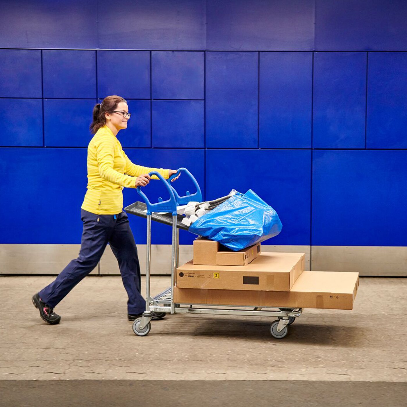 En IKEA medarbejder i gul trøje kører en fyldt IKEA kundevogn med tre pakker på på vejen foran en blå bygning.