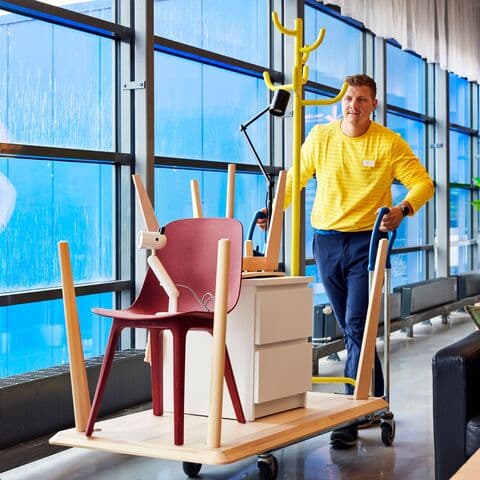En IKEA medarbejder i gul trøje transporterer en stol et bord og en hvid kommode på n vogn foran en række blå vinduer.