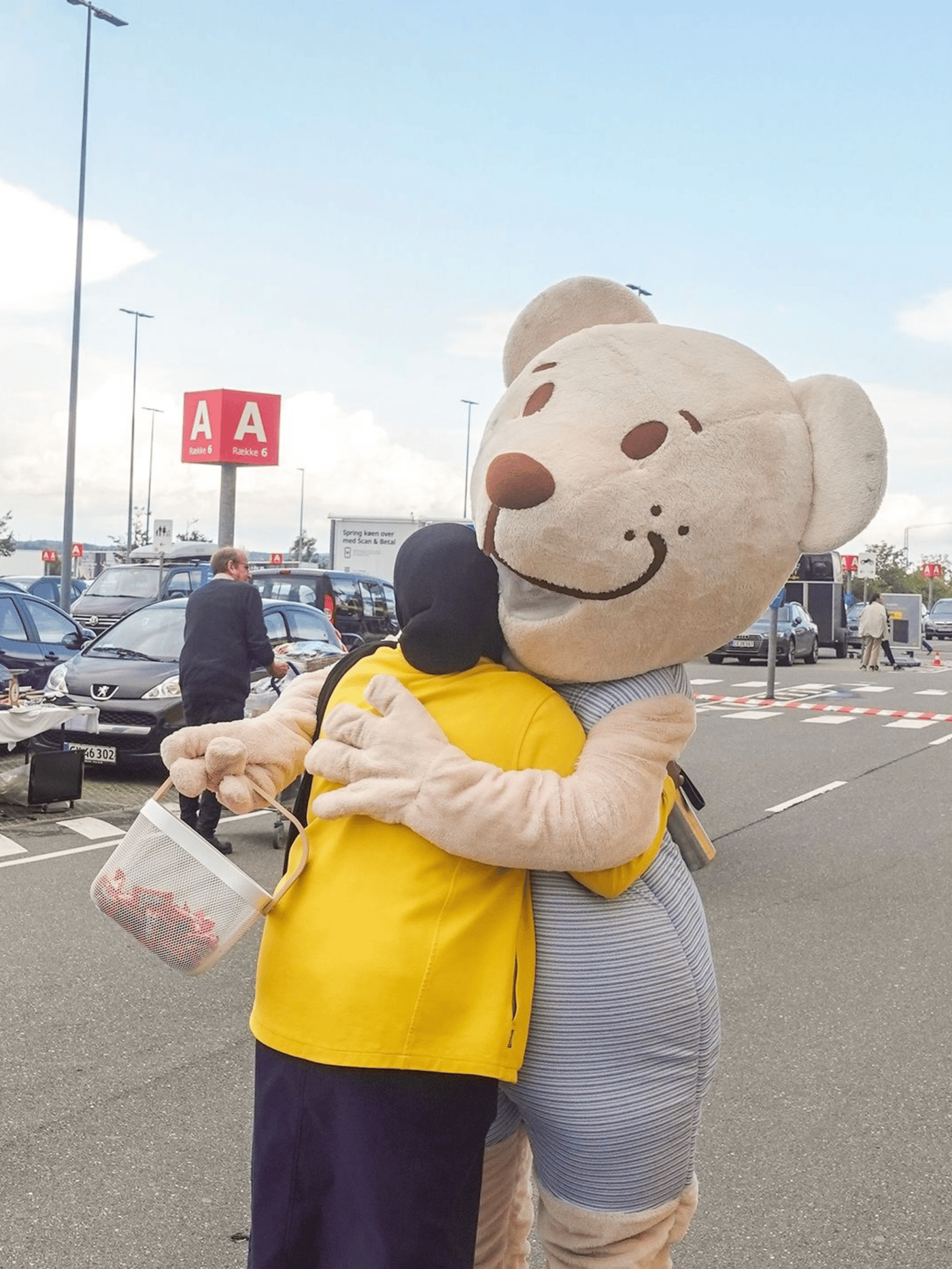 En IKEA medarbejder i sin uniform krammer maskotten Fabler Bjørn på en parkeringsplads.