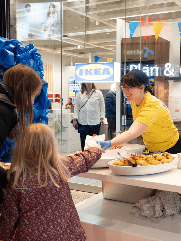 En IKEA medarbejder i sin uniform serverer kage til to børn ude foran IKEA Plan and Order Point Herning.