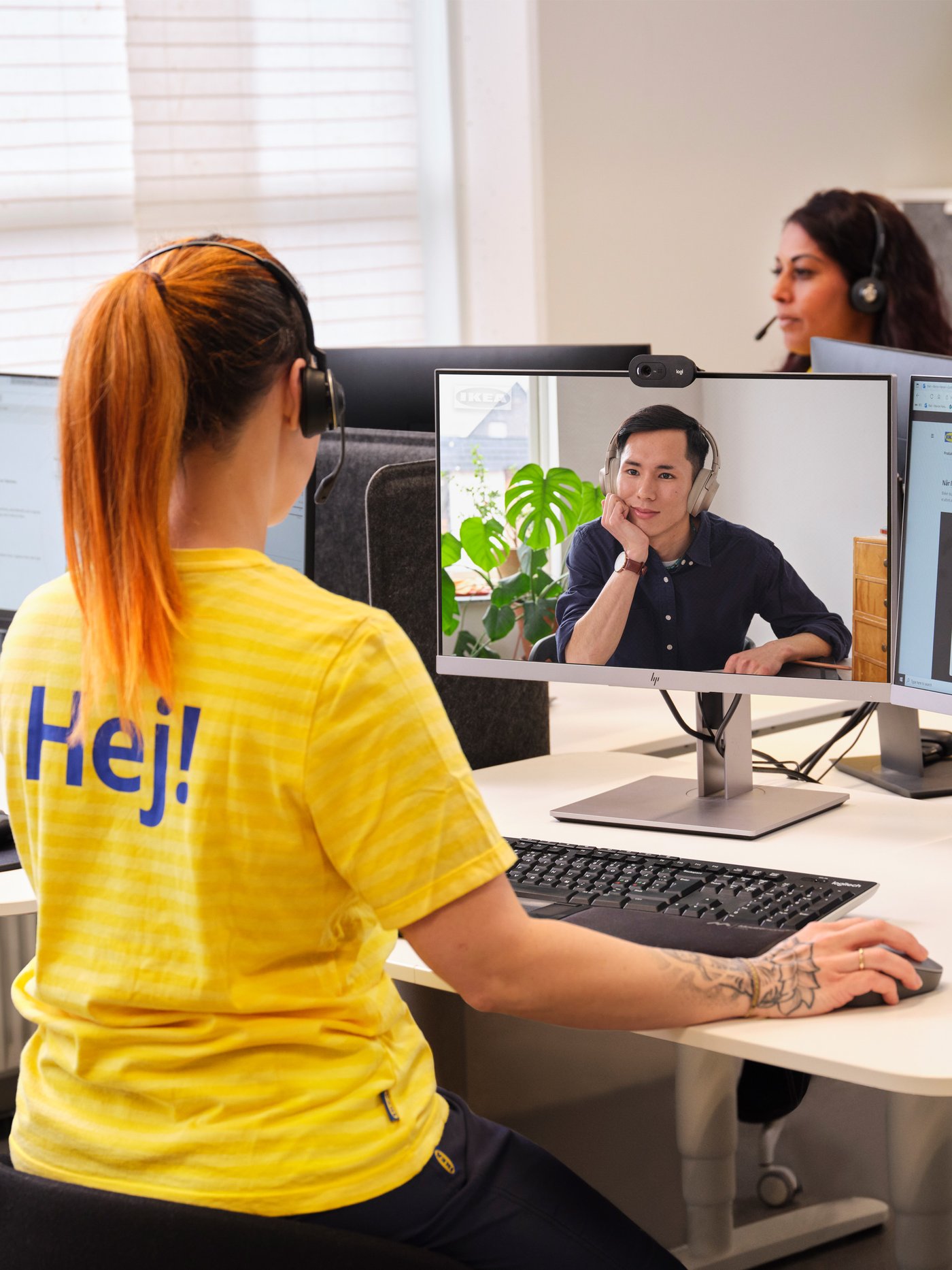 En IKEA medarbejder med en gul bluse hvor der står HEJ på står med ryggen til og kigger ind i en skærm hvor en mand sidder og lytter på hvad hun siger.