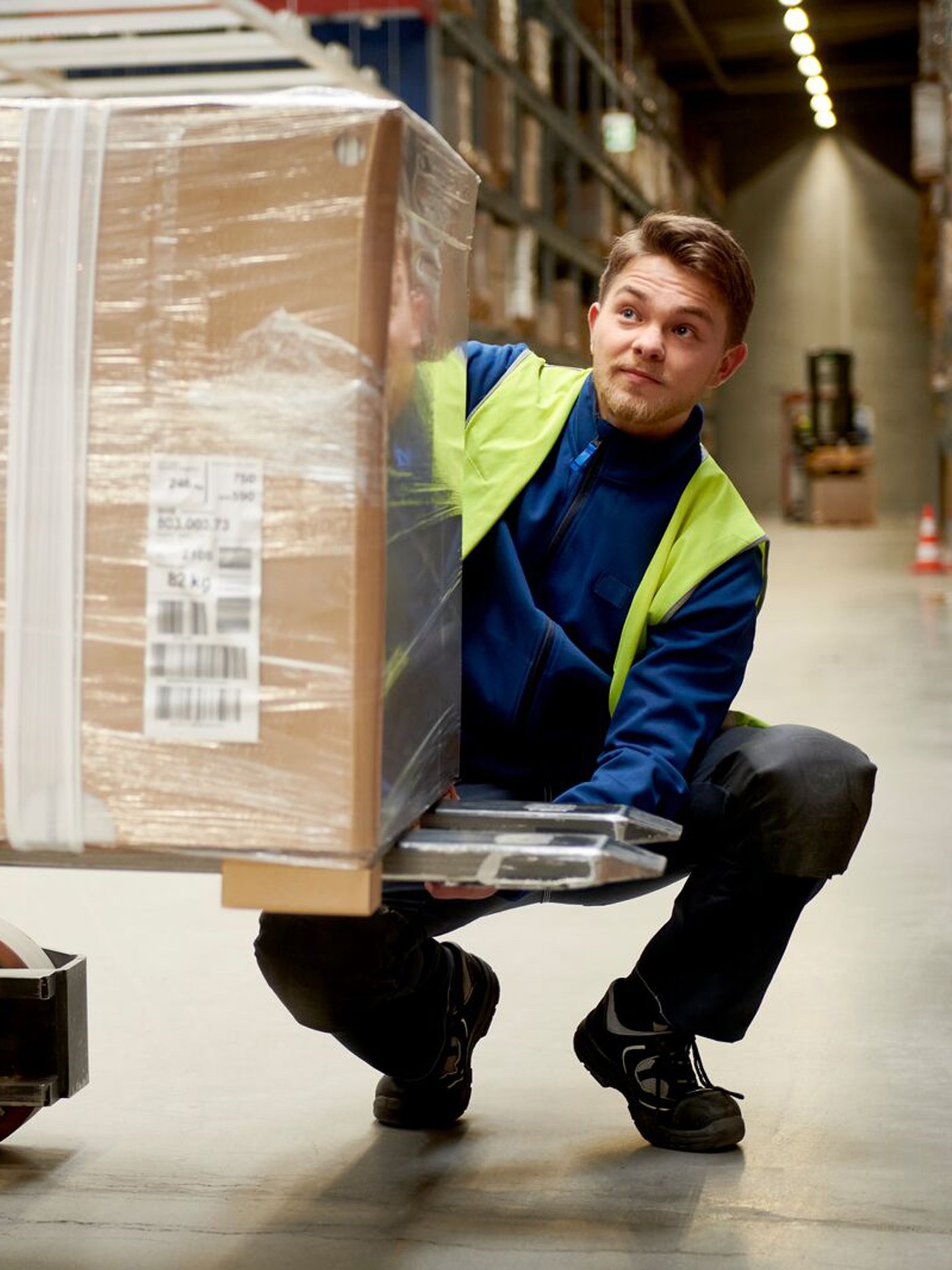 En IKEA medarbejder på et lager sidder på hug foran en palleløfter der bærer en pakke pakket ind i plastik.