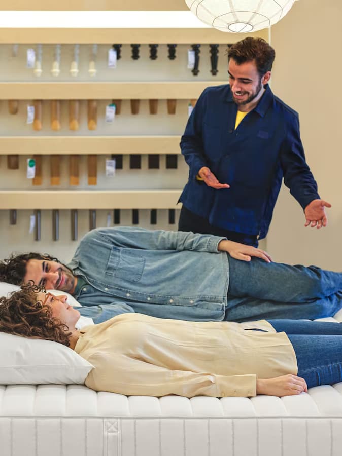 En IKEA medarbejder står i et vareus og gestikulerer med armene mens han viser en seng frem som et par kunder er ved at prøve ved at ligge ned i sengen.