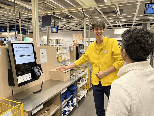 En IKEA medarbetare gestikulerar mot en självbetjäningskassa. I förgrunden syns en till person som tittar på medarbetaren.