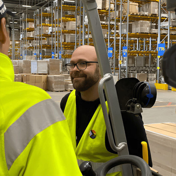 En IKEA medarbetare med säkerhetsväst och glasögon står vid en truck på ett lager. I förgrunden syns en annan medarbetare som sitter i trucken.