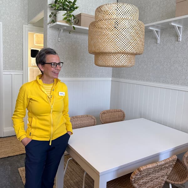 En IKEA medarbetare står i ett inrett rum med vitt bord, rottingstolar och lampa i naturmaterial på Stadsmissionen i Umeå.